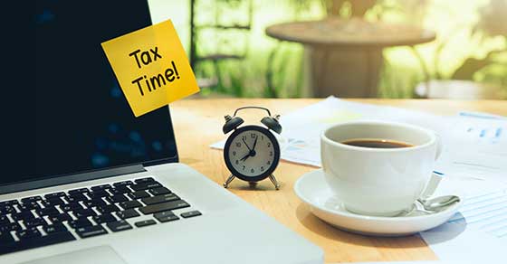 Laptop on desk, alarm clock, coffee cup, sticky note, Text: Tax Time,