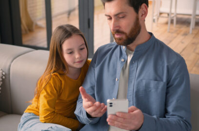 Adult man talking to his child on the couch, explaining something on the screen of his mobile phone