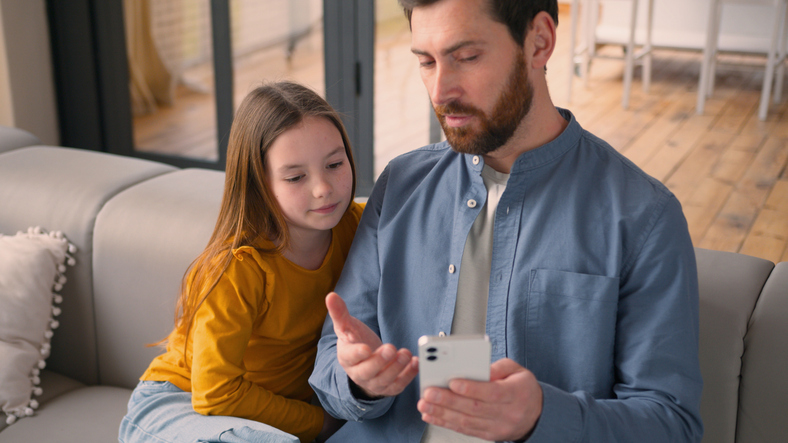 Adult man talking to his child on the couch, explaining something on the screen of his mobile phone