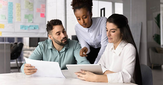 Three,Colleagues,Gathered,Around,Conference,Table,,Looking,At,Tablet,,Hold