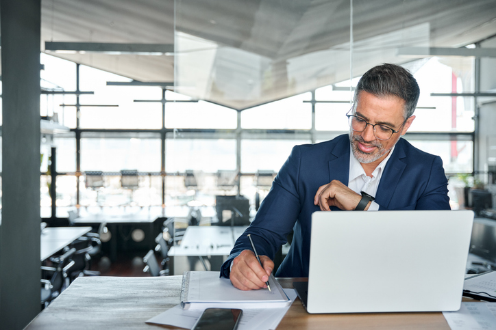 Middle-aged business man sitting at a desk at a corporate office building, sitting in front of laptop and taking notes in a notebook