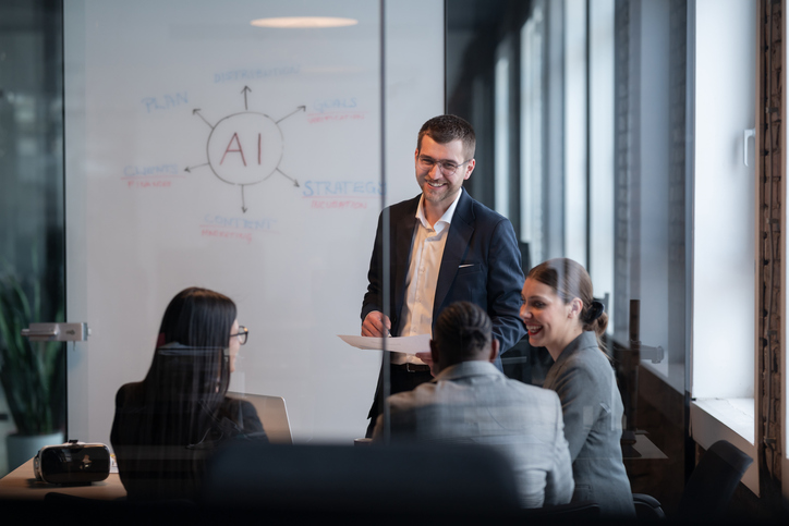 Team of four people in a modern office building, sitting around conference table in front of a whiteboard