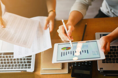 Close up of hands looking over papers, laptop, tablets featuring financial tables and charts