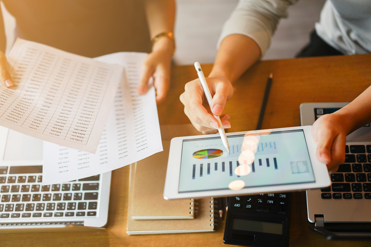 Close up of hands looking over papers, laptop, tablets featuring financial tables and charts