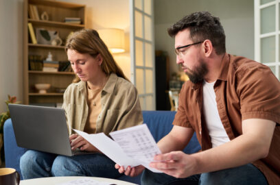 Young couple sitting on couch, man holding documents, woman looking down at laptop