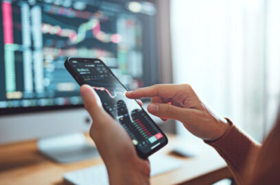 Closeup of someone looking at financial charts on their phone, computer in background