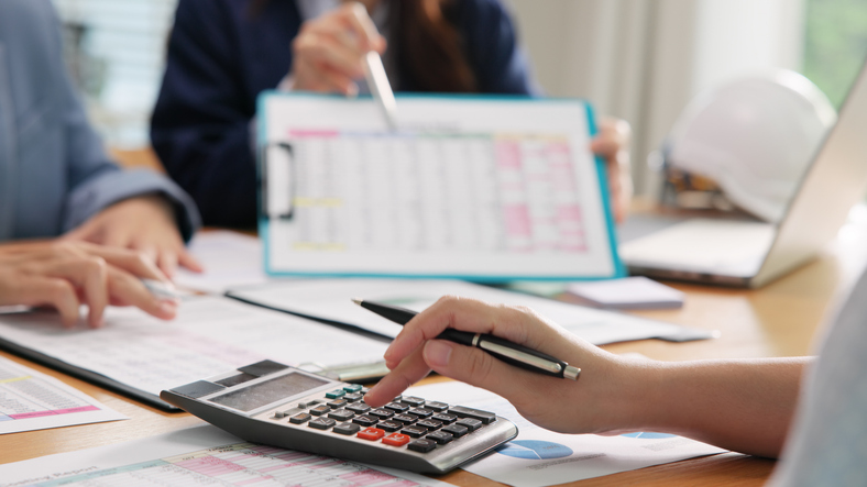 Closeup of conference table; people reviewing numbers in business meeting, financial tables and charts spread across conference table