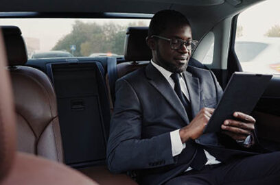 Smiling businessman using digital tablet, reading good news sitting on backseat of car. Handsome african american man in suit writing something down in personal organizer while sitting inside of car.