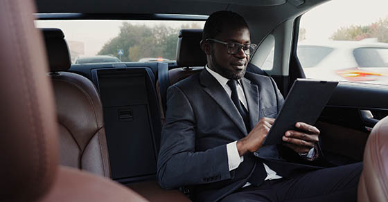 Smiling businessman using digital tablet, reading good news sitting on backseat of car. Handsome african american man in suit writing something down in personal organizer while sitting inside of car.
