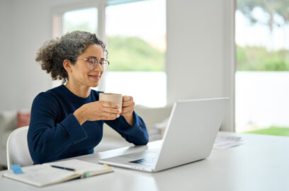 Woman sitting at home holding coffee and smiling at laptop, watching webinar in modern house