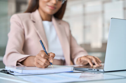 Close up view of professional business woman working on laptop computer writing notes in notebook at work in corporate office.