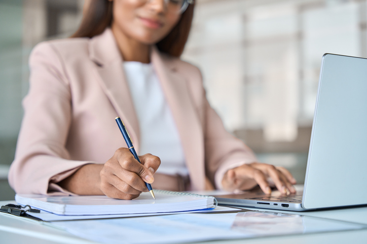 Close up view of professional business woman working on laptop computer writing notes in notebook at work in corporate office.