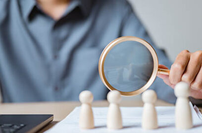 Close-up of a person using magnifying glass to examine wooden human figures, representing recruitment, HR management, candidate selection, and employee evaluation in business.