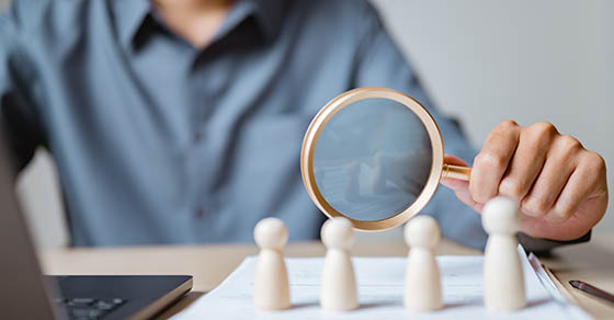 Close-up of a person using magnifying glass to examine wooden human figures, representing recruitment, HR management, candidate selection, and employee evaluation in business.