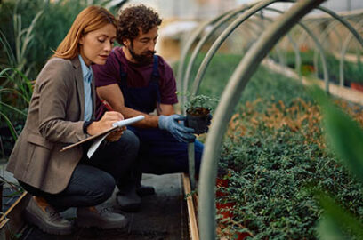 Businesswoman taking notes while examining quality of potted plants with a greenhouse worker.