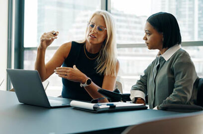 A mature woman and a young woman using a wheelchair cooperate on a project, emphasizing workplace collaboration.