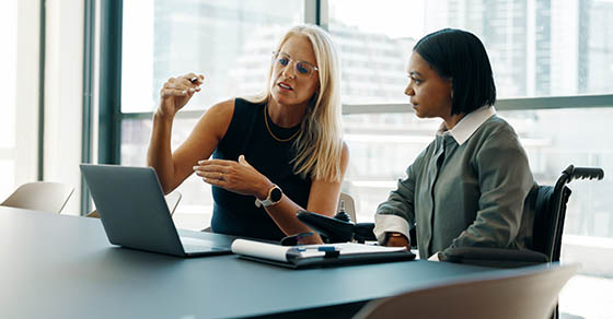 A mature woman and a young woman using a wheelchair cooperate on a project, emphasizing workplace collaboration.